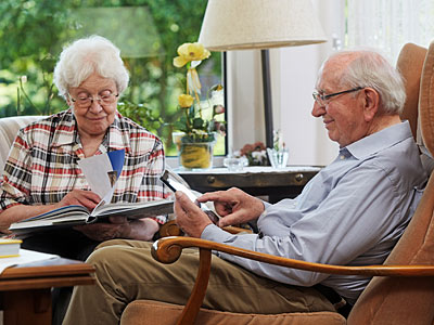 Ein älteres Paar sitzt in einem gemütlichen Raum. Die Frau liest ein Buch, während der Mann ein Tablet benutzt. Pflanzen und Dekoration sind im Hintergrund zu sehen.