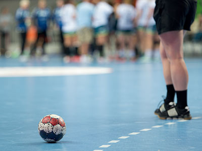 Ein Handball liegt auf dem Spielfeld, während Spieler im Hintergrund stehen.
